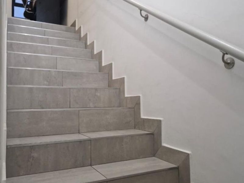 A staircase featuring modern grey tiled steps and a wall-mounted handrail, leading to an upper landing area.