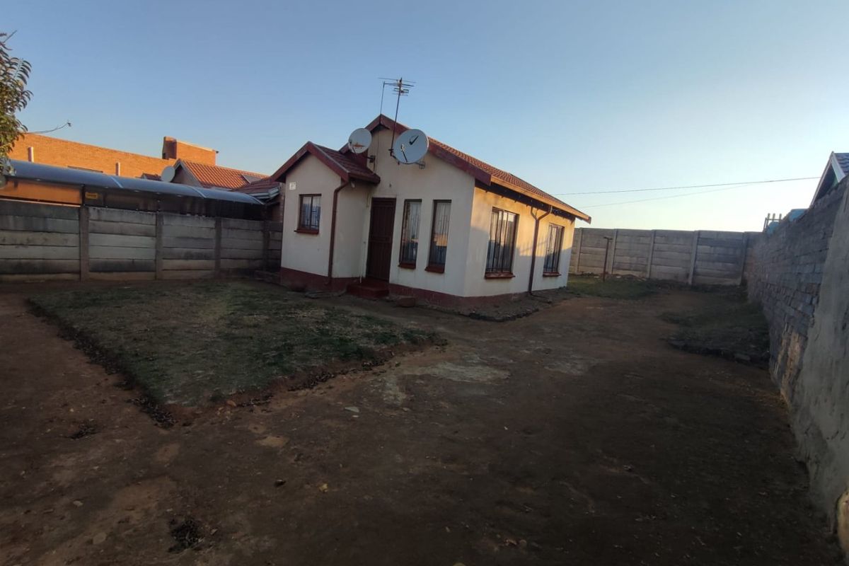 A detached single-story house featuring a tiled roof, security bars on windows, and a large, unpaved yard enclosed by perimeter walling.