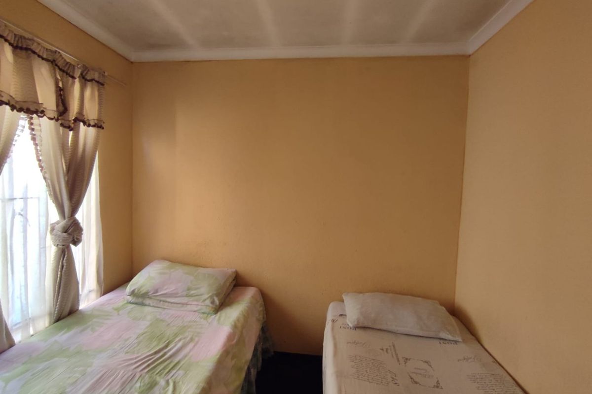 A compact bedroom featuring plain walls, a ceiling with visible staining, and a window with curtains.