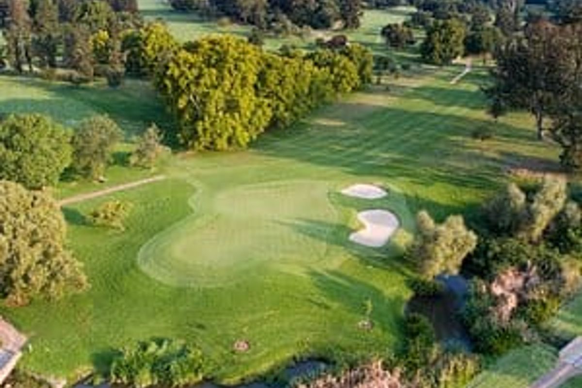 An aerial view of a residential vacant land plot situated within a lush, green golf course estate featuring mature trees and a small water feature.