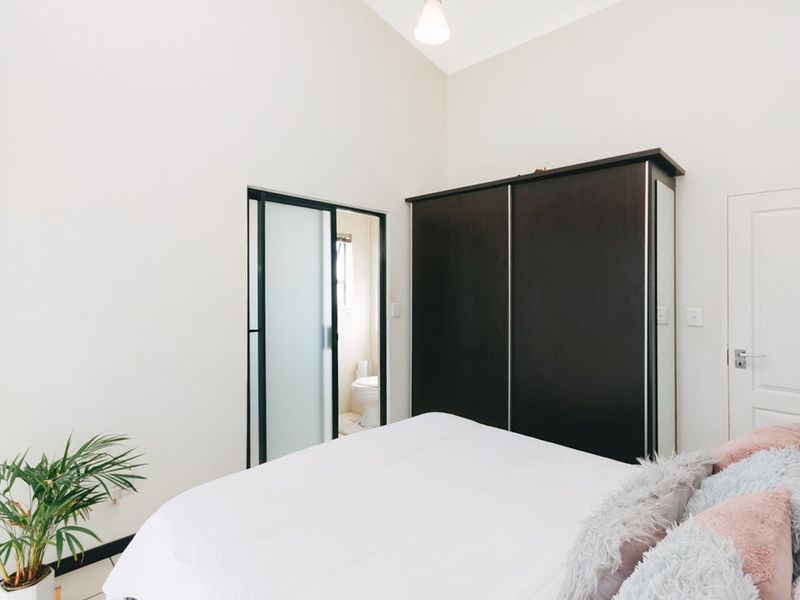 A bright bedroom featuring high ceilings, a built-in dark wood wardrobe, and an en-suite bathroom accessible through a modern sliding glass door.