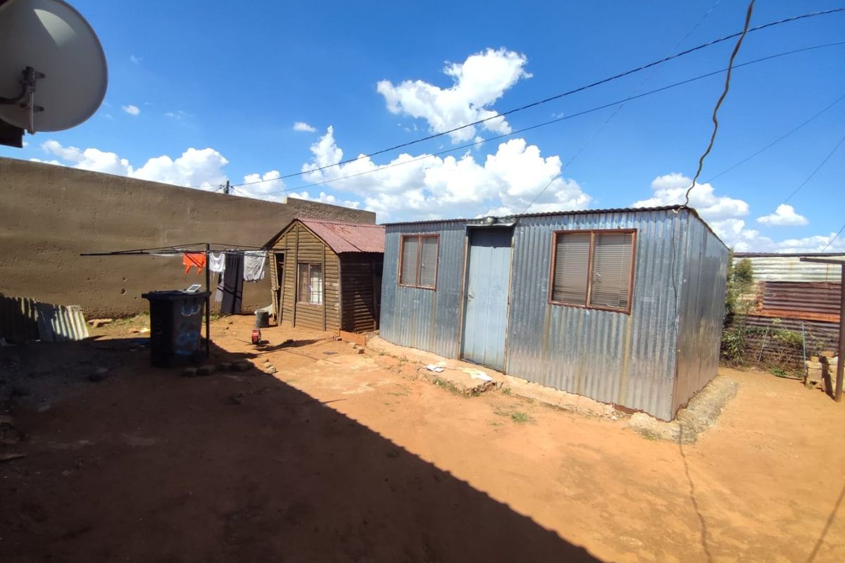 Exterior view of a corrugated metal structure and a small wooden outbuilding situated on an unpaved plot.