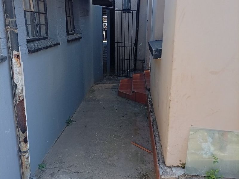 A narrow exterior side passage between two building structures featuring concrete paving, a metal security gate, and a rusted drainage downpipe.