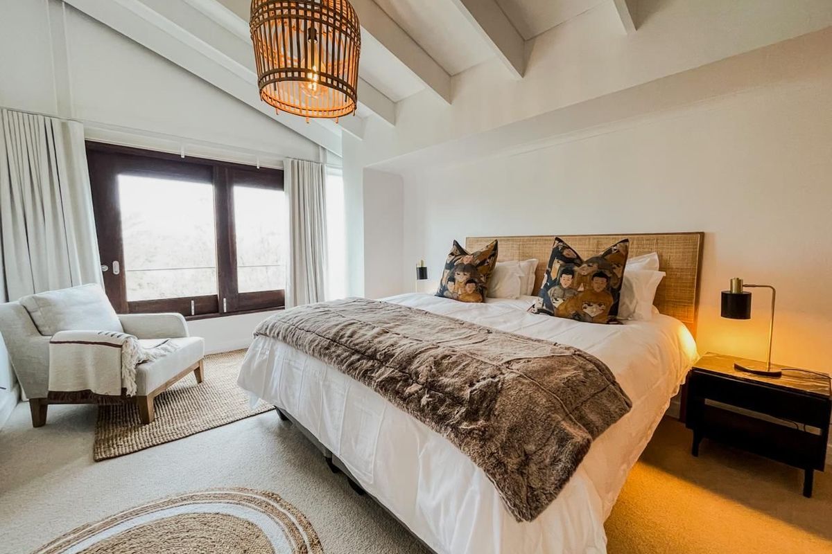 A well-lit bedroom featuring vaulted ceilings with exposed beams, neutral carpeting, and large windows providing natural light.