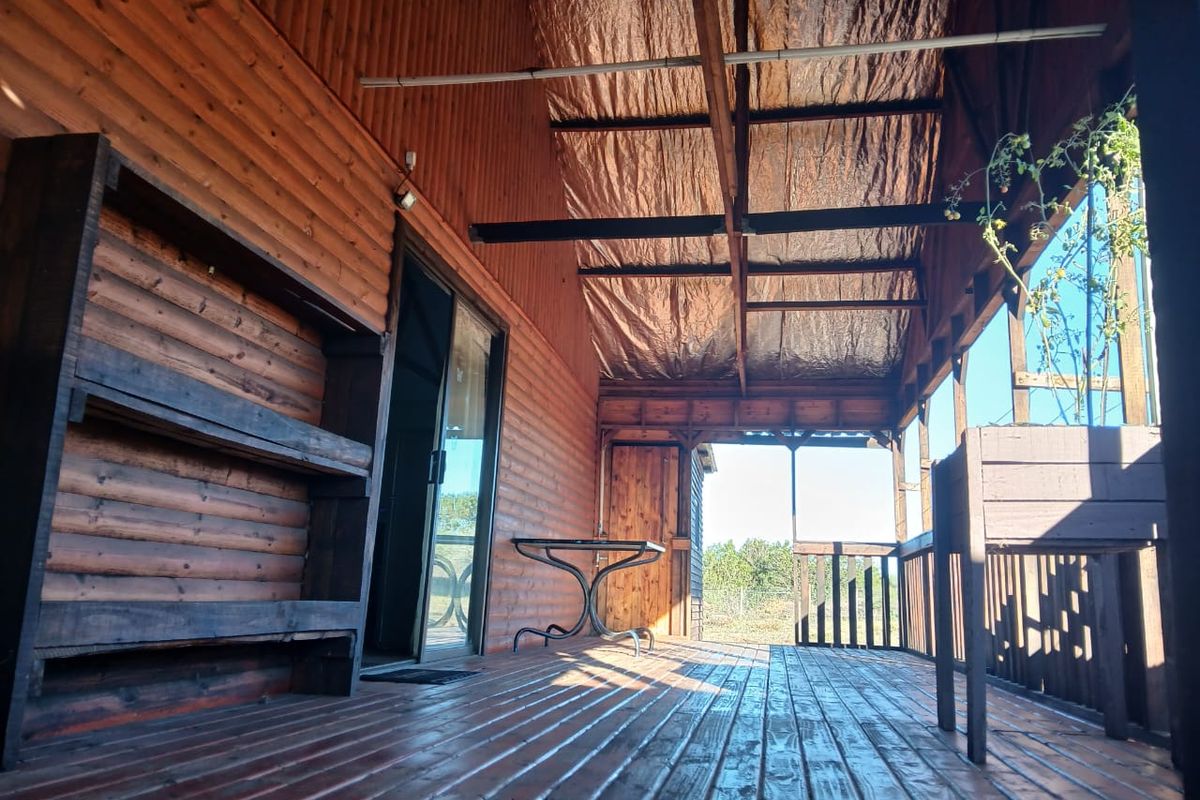 A covered timber deck featuring wooden wall cladding, a sliding glass door entrance, and open-air views, providing a rustic outdoor living space.