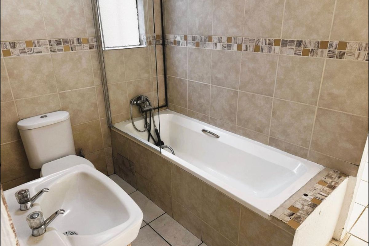 A functional bathroom featuring a built-in bathtub with a glass shower screen, a pedestal basin, and a toilet, finished with neutral wall and floor tiling.