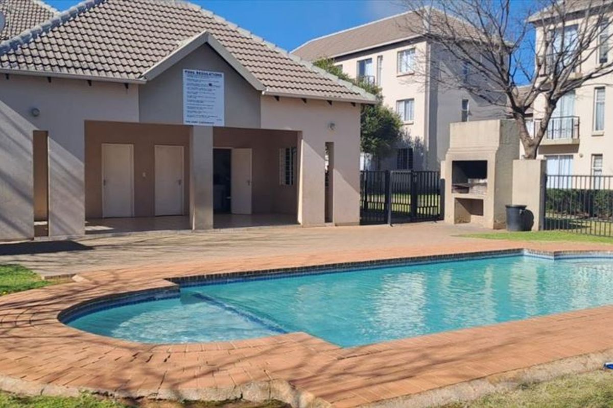 Communal swimming pool area featuring a paved deck, built-in braai, and a covered clubhouse structure with tiled roof.