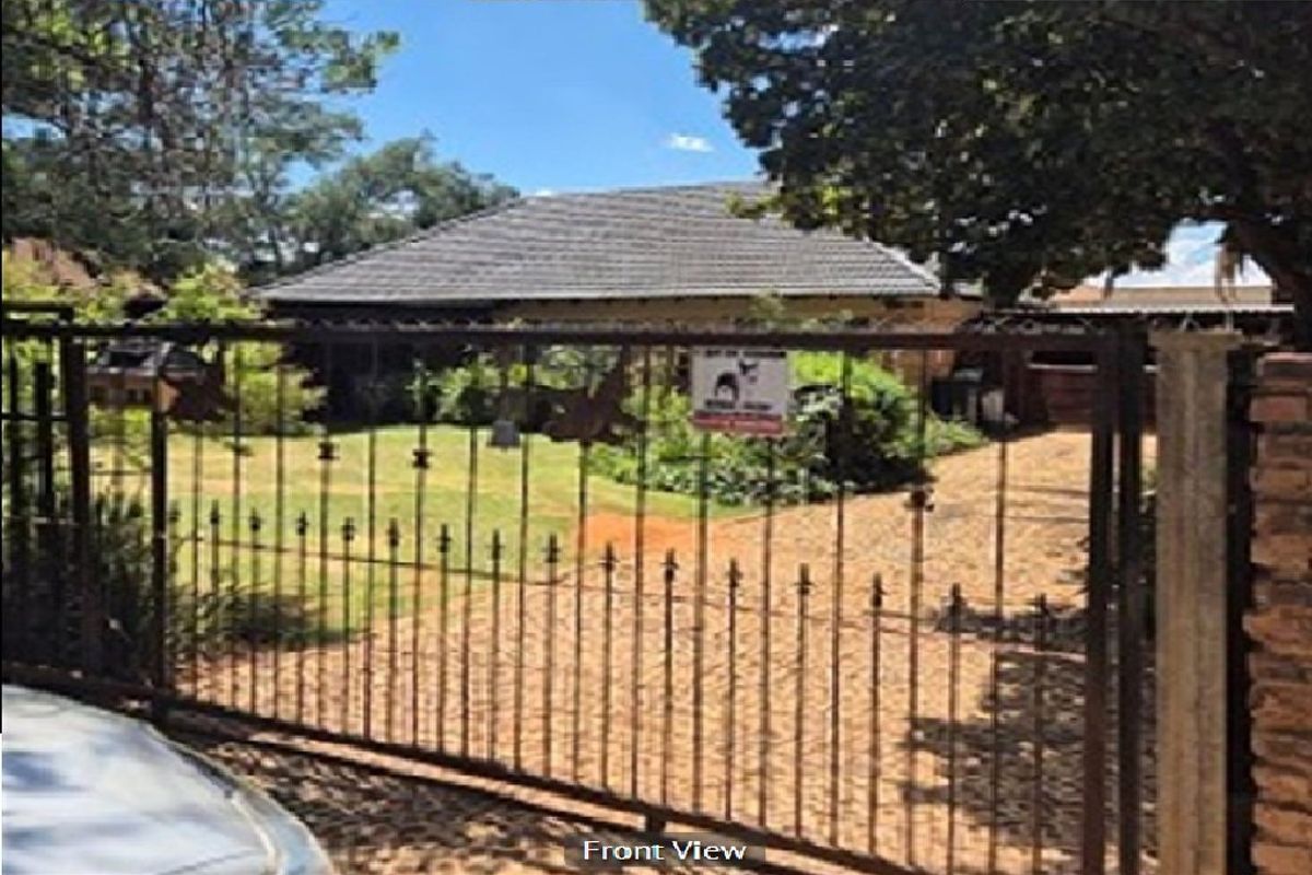 Front view of the property featuring a metal security gate, paved driveway, and established garden leading to the main house entrance.