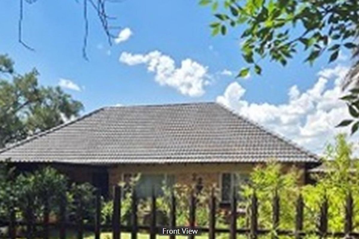Front view of a single-story residential house featuring a tiled roof and brick facade, partially obscured by mature garden foliage and a perimeter fence.