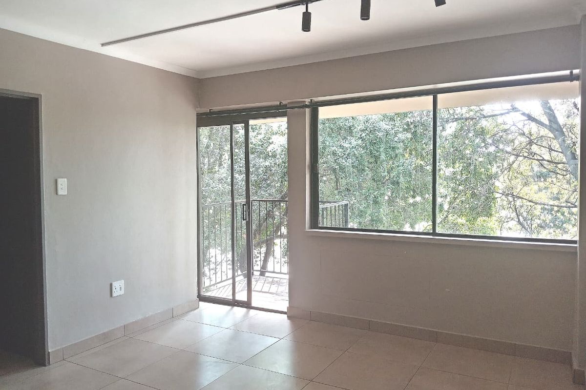A bright, empty living area featuring neutral tiled flooring, track lighting, and large windows with access to a small balcony.