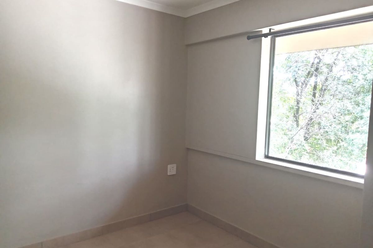 A bright, empty bedroom featuring neutral wall tones, tiled flooring, and a large window providing natural light and a view of the surrounding trees.