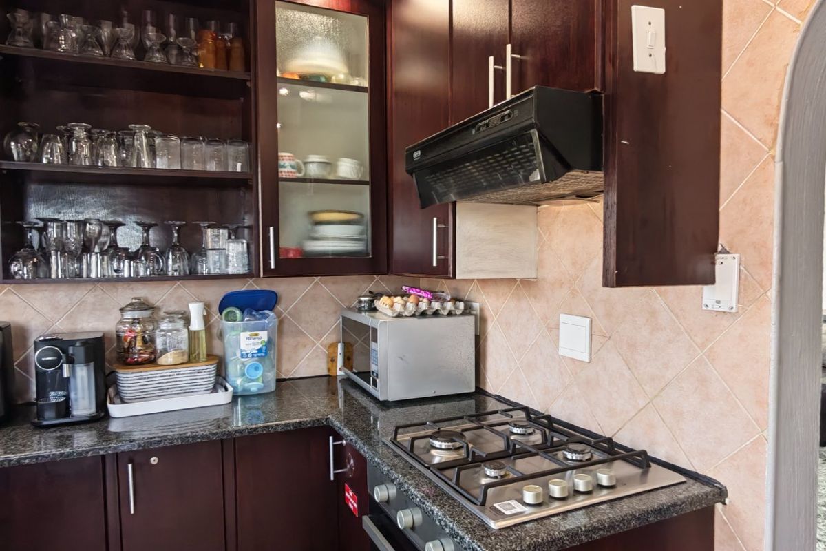 A functional kitchen featuring dark wood cabinetry, granite countertops, a gas hob with extractor fan, and a tiled backsplash, leading into an adjacent living area.