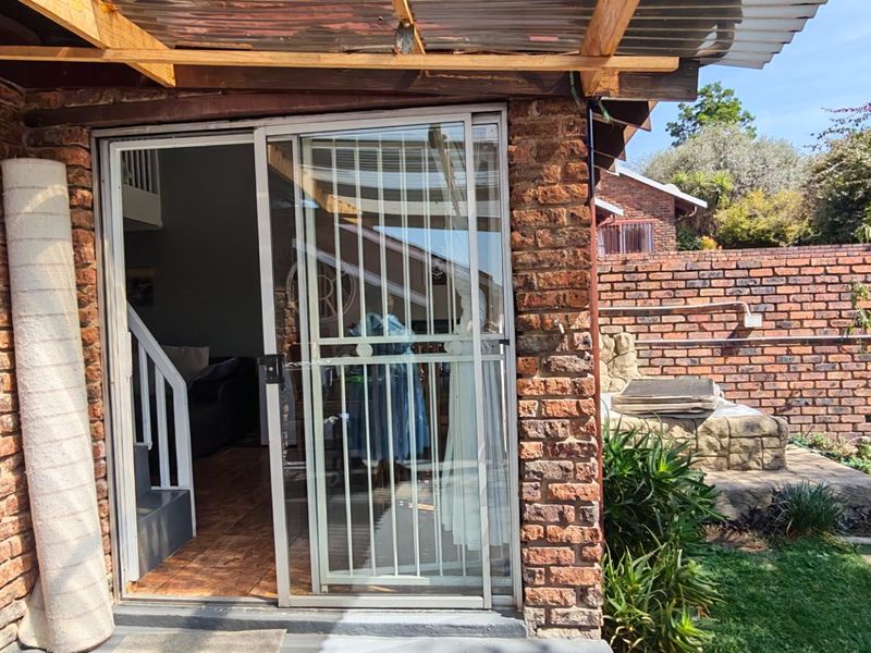 Rear patio area featuring a brick facade, sliding glass door with security gate, and a timber-framed corrugated roof overlooking a small garden.