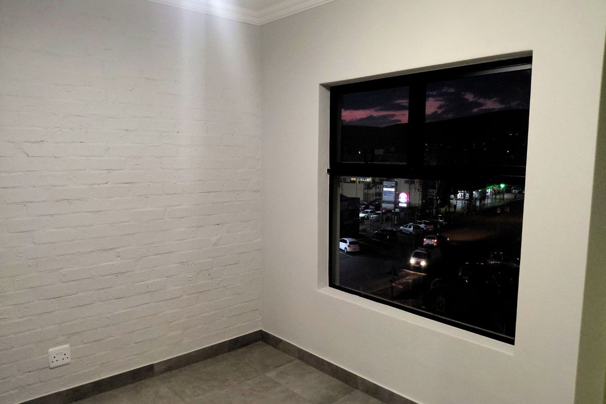 A clean, empty bedroom featuring a painted brick feature wall, tiled flooring, and a large window with views of the street at dusk.