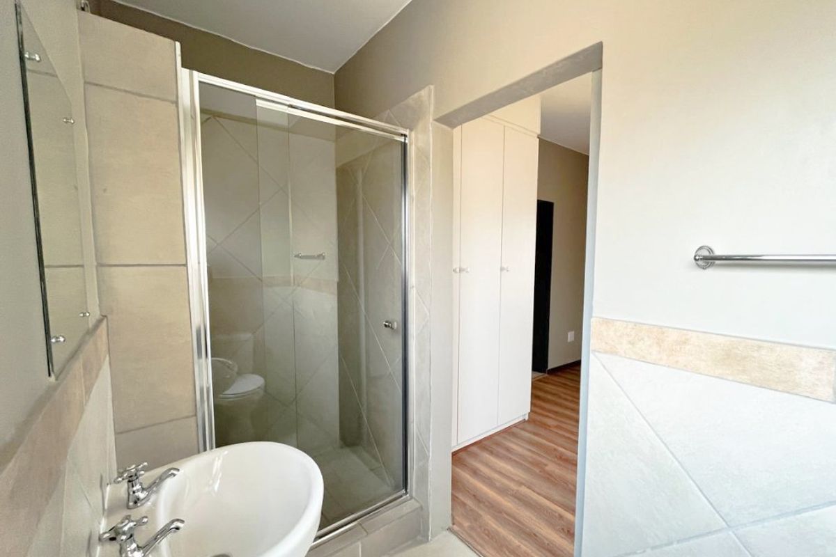 A clean, functional bathroom featuring a glass shower enclosure, pedestal basin, and neutral wall tiling, with adjacent built-in storage cupboards.
