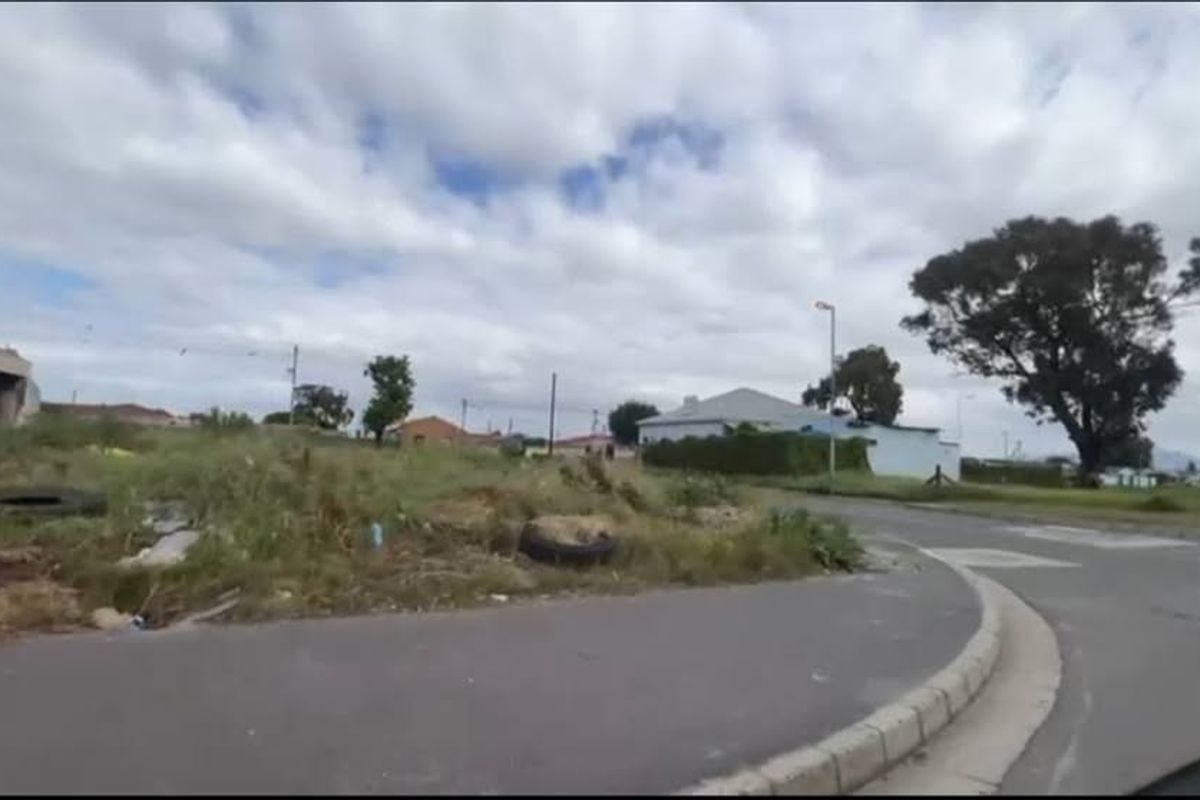 A street-level view of a vacant residential plot featuring asphalt road access, concrete curbing, and surrounding neighborhood infrastructure.