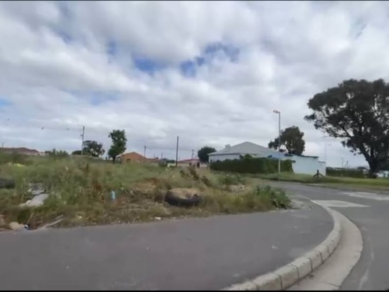 A street-level view of a vacant residential plot featuring asphalt road access, concrete curbing, and surrounding neighborhood infrastructure.