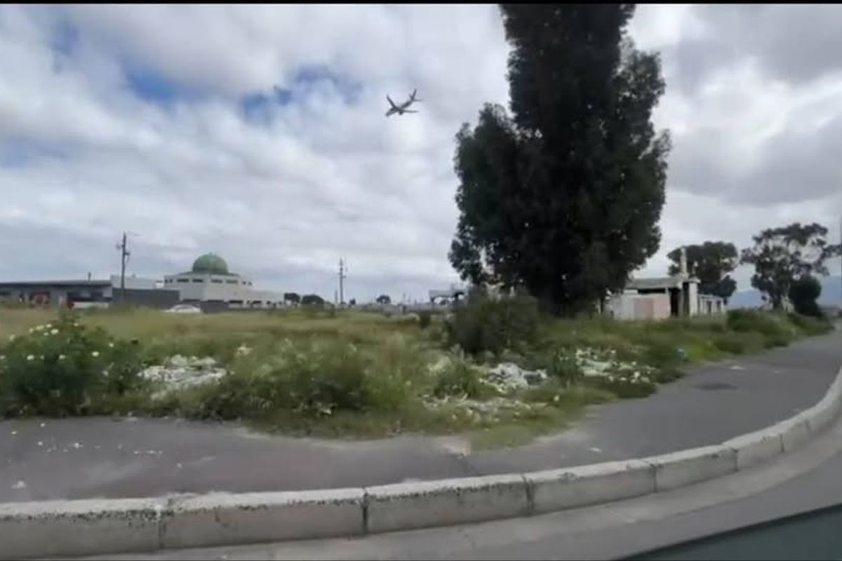 A street-level view of a vacant residential plot featuring overgrown vegetation and debris, situated near established infrastructure and local amenities.
