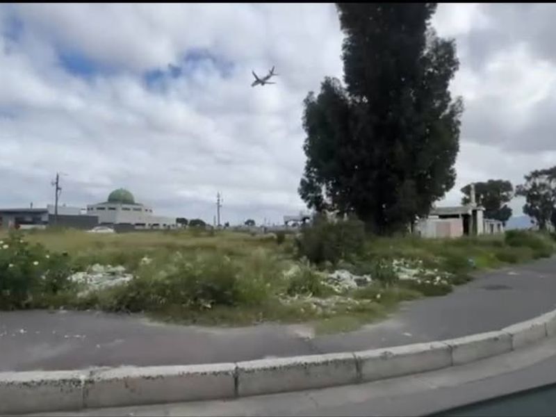 A street-level view of a vacant residential plot featuring overgrown vegetation and debris, situated near established infrastructure and local amenities.