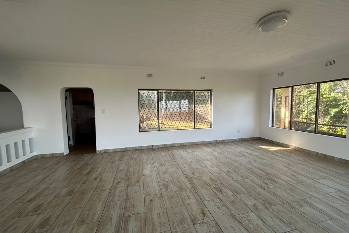 A bright, empty living room featuring wood-look tile flooring, large windows with security bars, and a classic tongue and groove ceiling.
