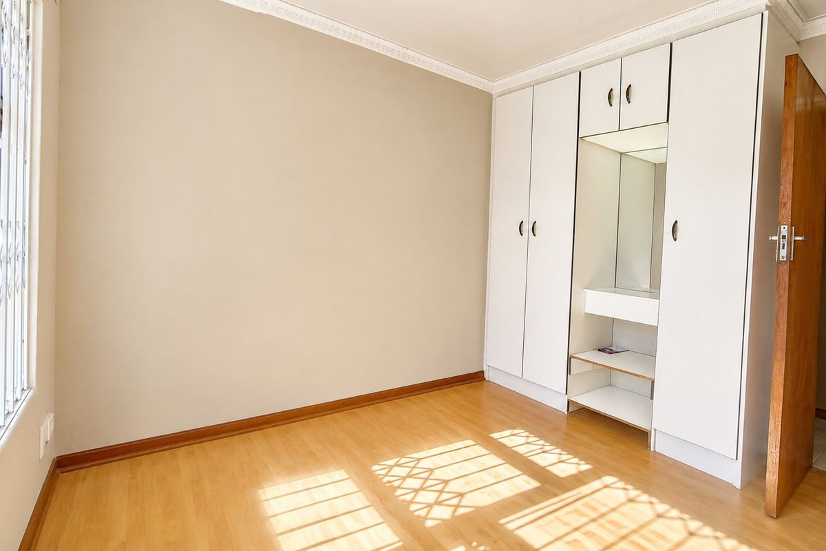 A bright, empty bedroom featuring light laminate flooring, built-in cupboards with a central vanity mirror, and a security gate on the window.