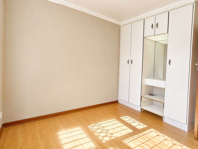 A bright, empty bedroom featuring light laminate flooring, built-in cupboards with a central vanity mirror, and a security gate on the window.