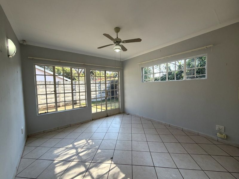 A bright, empty living room featuring tiled flooring, a ceiling fan, and large grid-pattern windows providing natural light and garden access.
