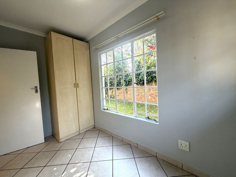 A bright, empty bedroom featuring tiled flooring, a built-in wardrobe, and a large grid-pattern window overlooking the garden.