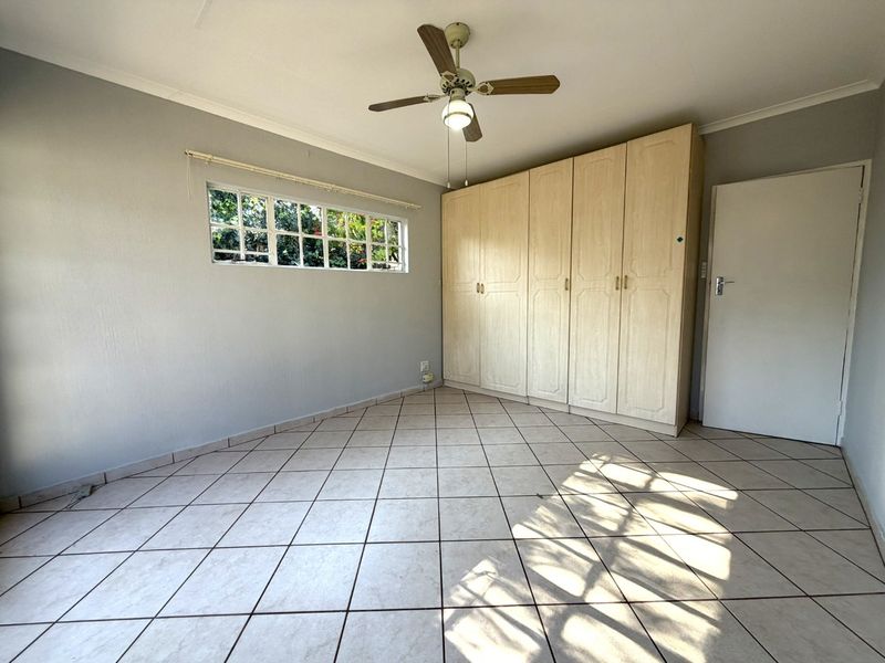 A spacious, empty bedroom featuring neutral tiled flooring, built-in wooden cupboards, a ceiling fan, and a horizontal window providing natural light.