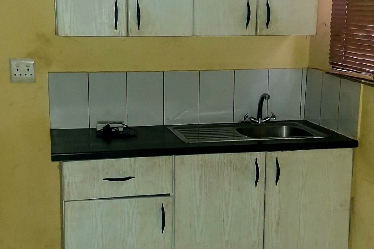 A compact kitchen area featuring basic laminate cabinetry, a stainless steel sink, tiled splashback, and ceramic floor tiles, requiring cosmetic updates.