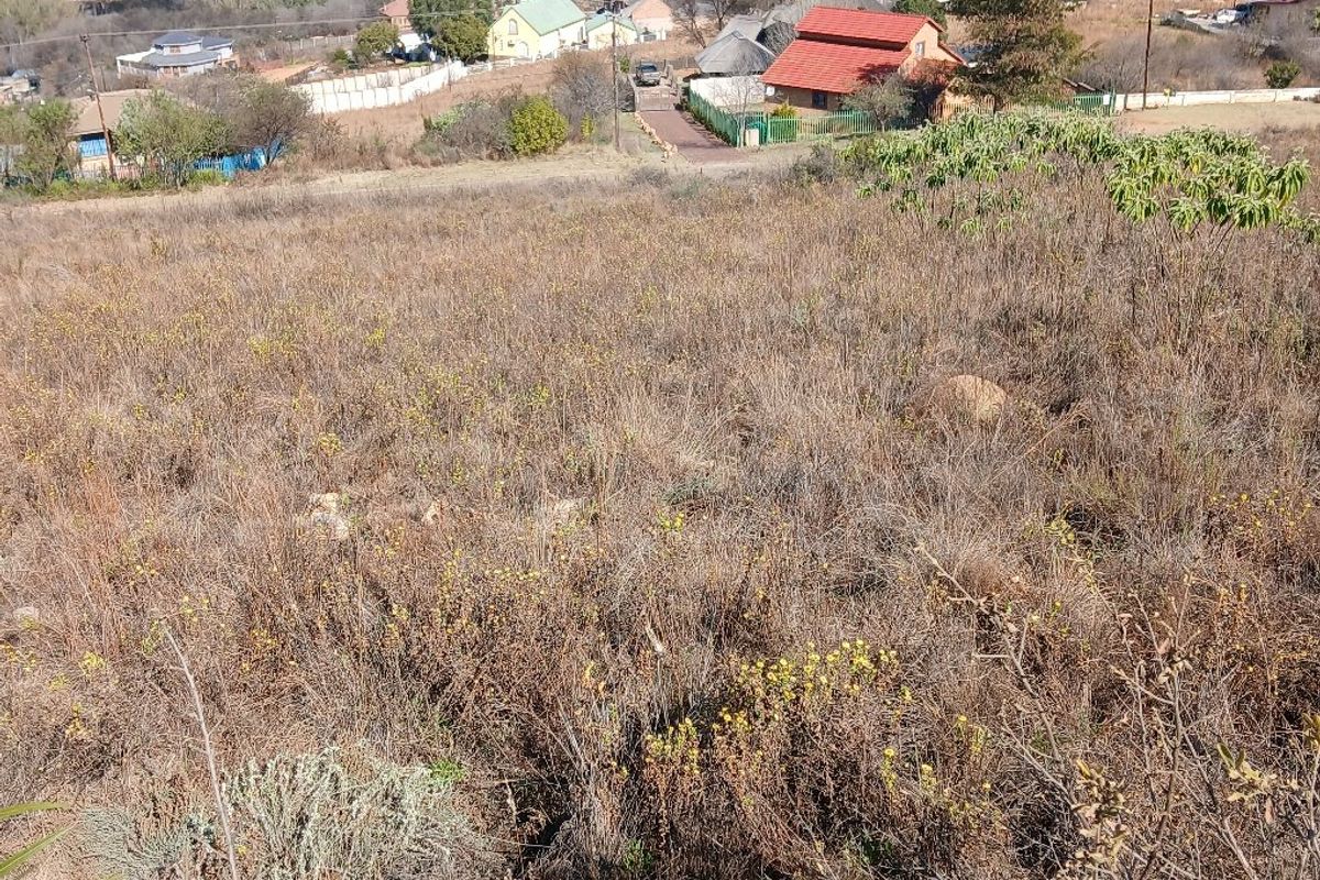 An elevated view of a sloped, undeveloped residential plot with surrounding neighborhood infrastructure visible in the background.