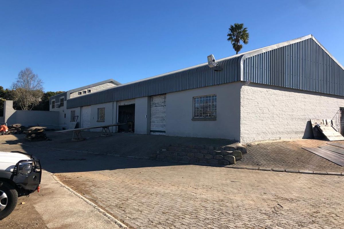 Exterior view of an industrial warehouse facility featuring corrugated metal roofing, roller shutter access doors, and a paved driveway area.