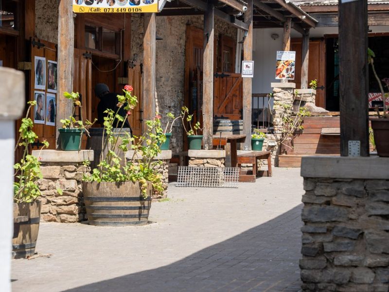 A commercial courtyard area featuring stone pillars, timber pergola structures, and paved walkways, providing access to various shop fronts.