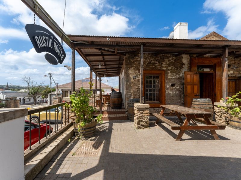A rustic outdoor patio area featuring stone masonry walls, a timber-framed veranda with a corrugated metal roof, and paved flooring.