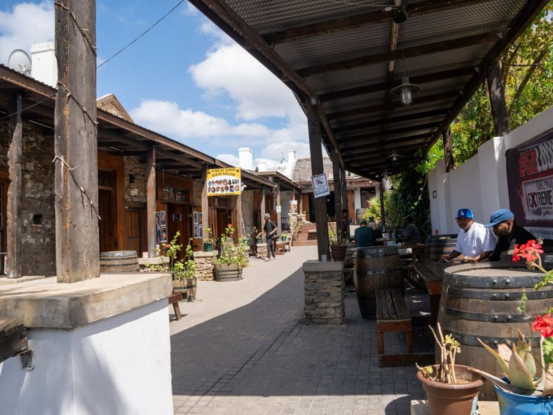 A rustic outdoor commercial courtyard featuring a covered walkway with timber beams, stone pillars, and paved flooring, providing a charming communal space.