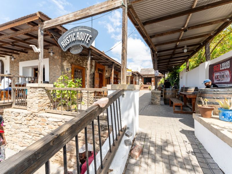 A rustic outdoor commercial patio area featuring stone masonry walls, timber pergola roofing, and paved walkways, providing a charming, open-air seating environment.