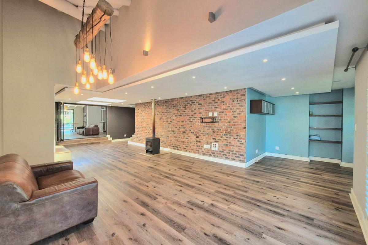 Spacious living area featuring a striking exposed brick wall, a modern freestanding fireplace, vaulted ceilings, and warm laminate flooring with integrated lighting.