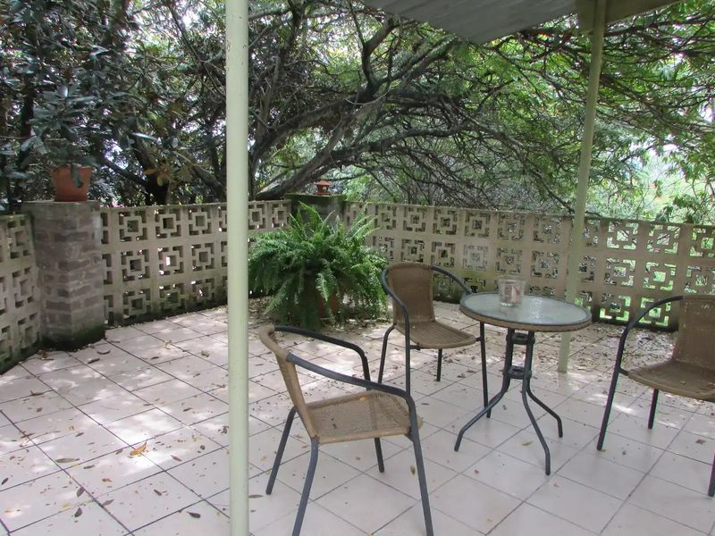A covered outdoor patio area featuring tiled flooring and a decorative breeze block perimeter wall, shaded by mature trees.