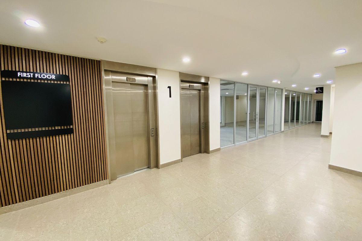 Modern commercial hallway featuring stainless steel elevators, timber slat wall detailing, and glass-partitioned office spaces with bright recessed lighting.