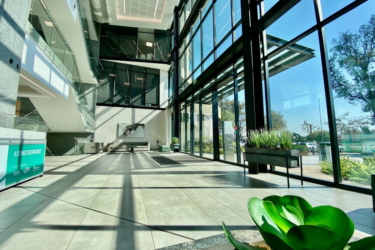 Modern commercial building entrance lobby featuring expansive floor-to-ceiling glass windows, high ceilings, and sleek tiled flooring with a contemporary staircase.