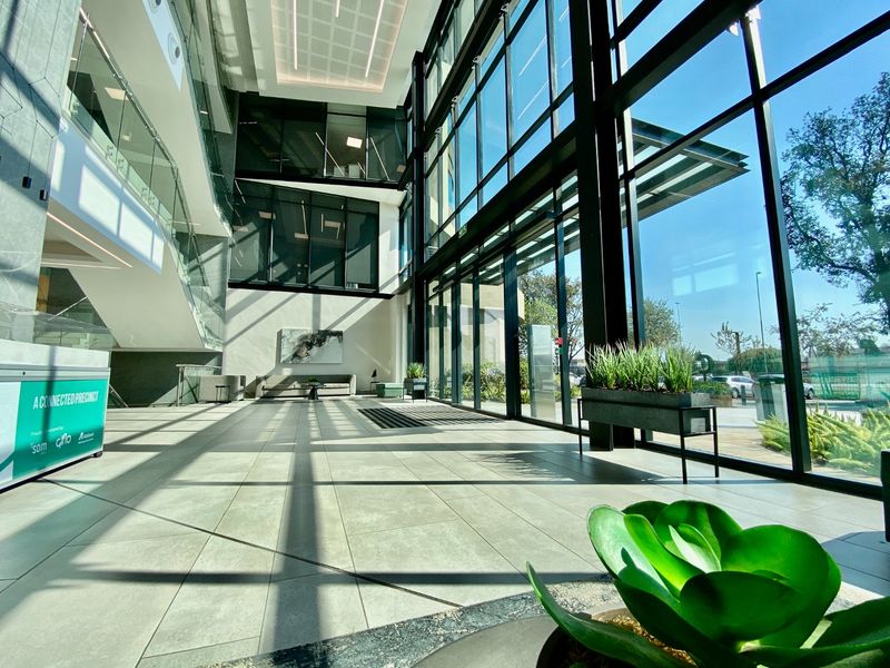 Modern commercial building entrance lobby featuring expansive floor-to-ceiling glass windows, high ceilings, and sleek tiled flooring with a contemporary staircase.