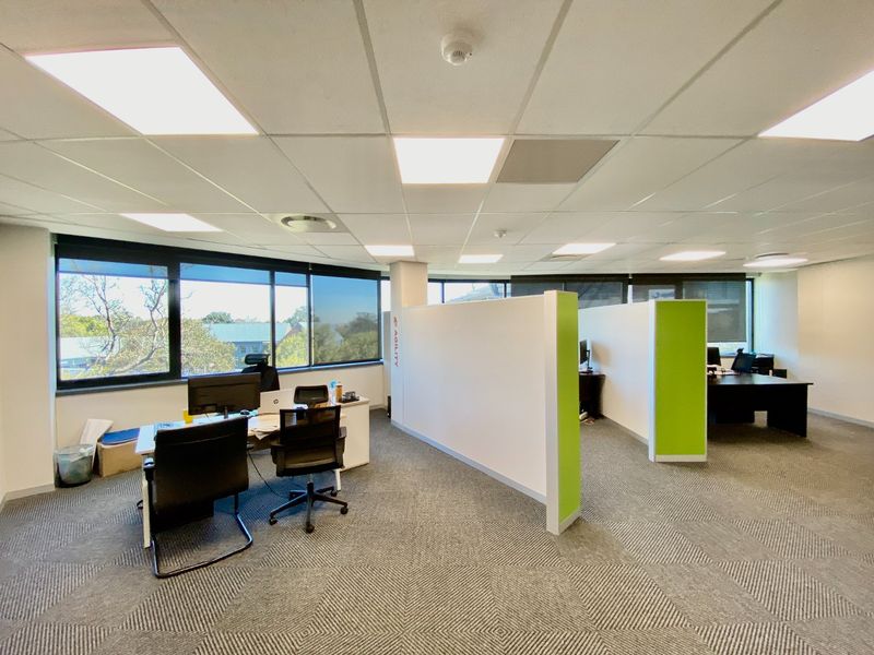 Modern open-plan office space featuring partitioned workstations, suspended ceilings with recessed lighting, carpeted flooring, and large windows providing ample natural light.