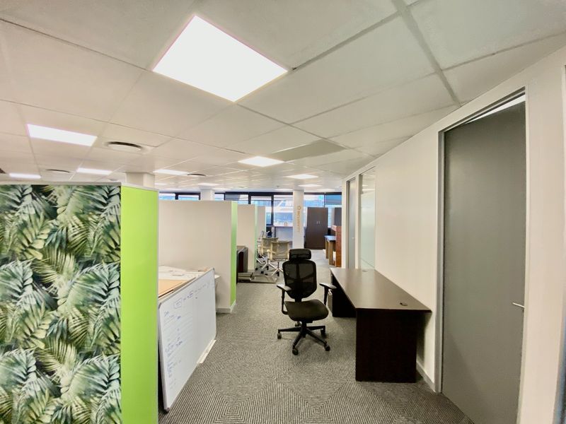 A modern open-plan office space featuring suspended ceilings, recessed lighting, carpeted flooring, and partitioned work areas with glass door access.