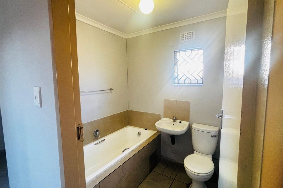A functional bathroom featuring a bathtub, wall-mounted basin, and toilet with neutral tiled finishes and a security-grille window.