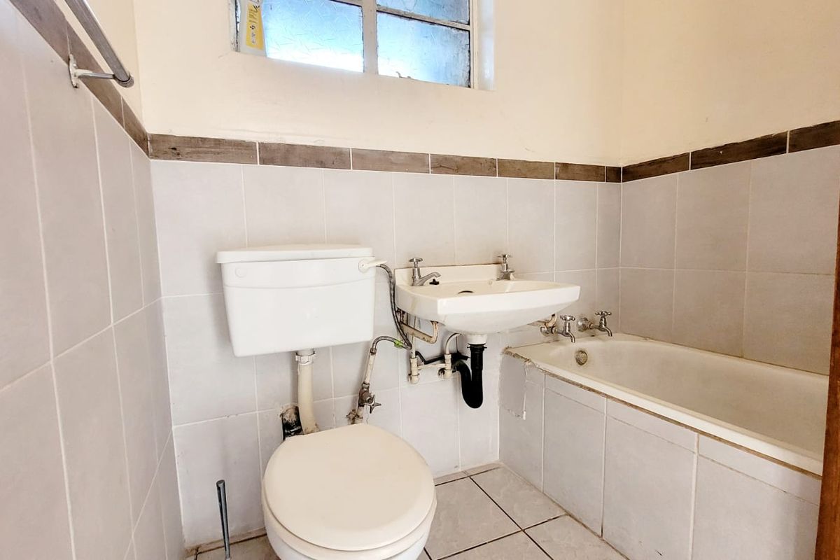 A compact bathroom featuring a standard toilet, wall-mounted basin, and bathtub with tiled finishes, requiring some cosmetic updates to the fixtures and tiling.