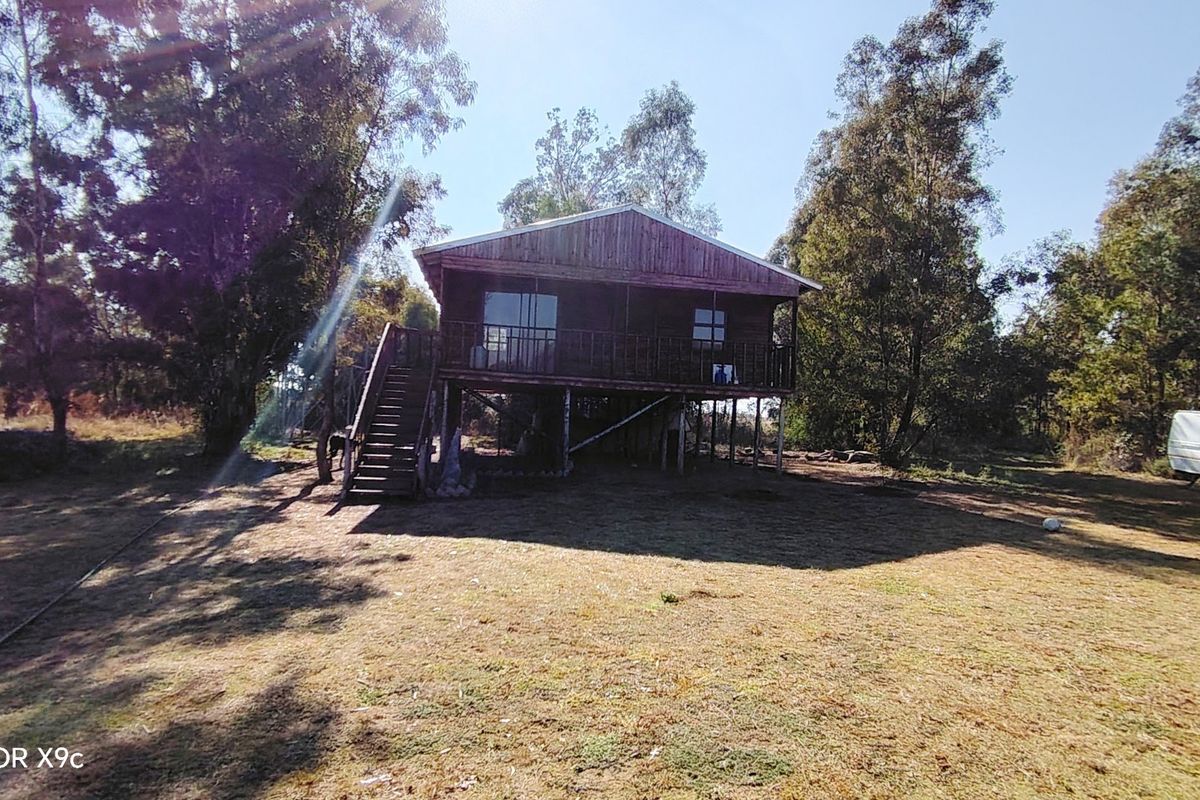 An elevated timber farm cottage featuring a staircase, balcony, and sliding glass doors, set within a rural landscape.