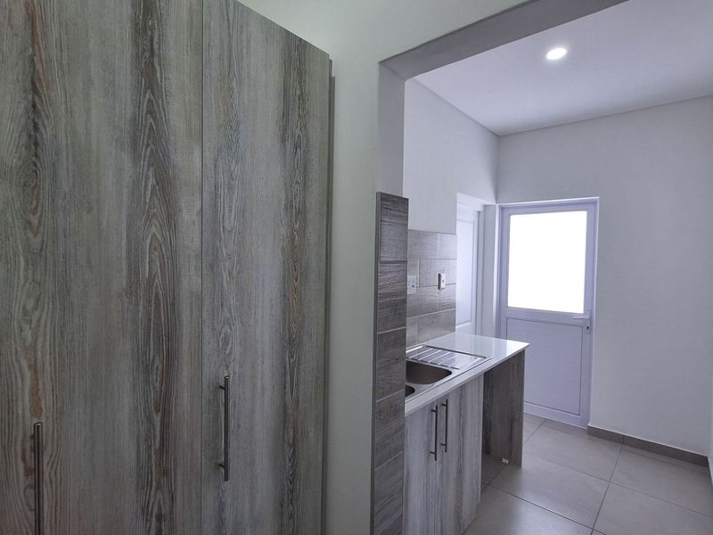 A clean, modern scullery featuring wood-look built-in cabinetry, a stainless steel sink, tiled splashback, and direct access to the exterior.