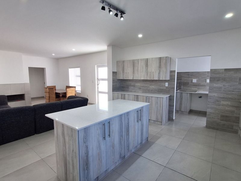 Spacious open-plan living area featuring a modern kitchen with a central island, stone countertops, and an adjoining scullery, finished with neutral floor tiles.