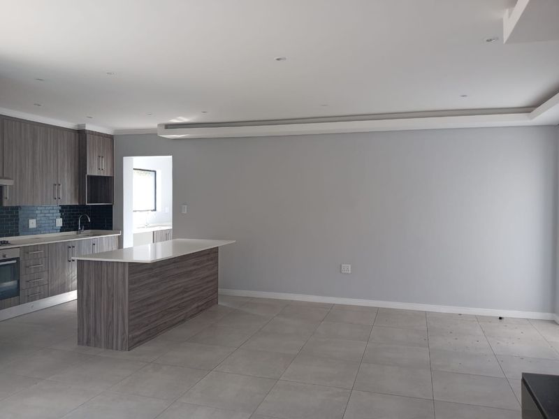 Modern open-plan living area featuring a contemporary kitchen with wood-grain cabinetry, stone countertops, built-in appliances, and neutral tiled flooring throughout.
