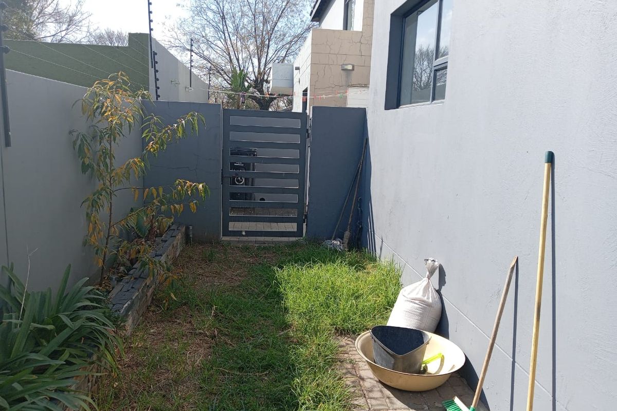 A narrow rear exterior side passage featuring a metal gate, paved walkway, and perimeter wall with electric fencing.